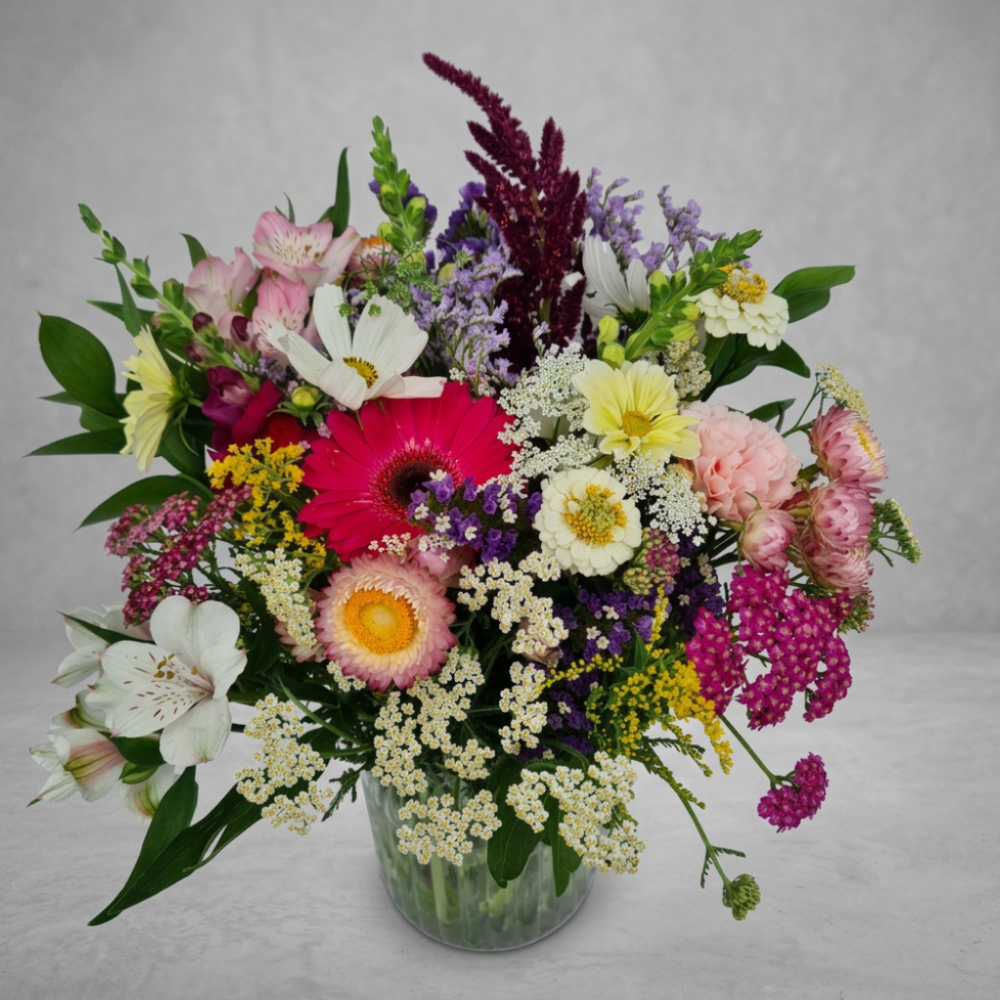 A vibrant and diverse seasonal summer bouquet, as shown in Seasonal Bouquet.png, featuring a colorful mix of flowers like hot pink gerbera daisies, white and yellow alstroemeria, strawflowers, amaranth, yarrow, and other wildflowers in a clear glass vase against a grey background.
