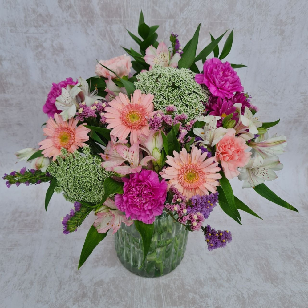 A bright and festive hand-tied bouquet featuring a vibrant mix of Guernsey-grown flowers, including bold yellow and orange freesias, deep pink heirloom carnations, and purple accents, finished with fresh green foliage.